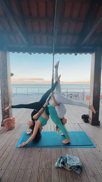People practising aerial yoga together on a covered wooden terrace beside the sea